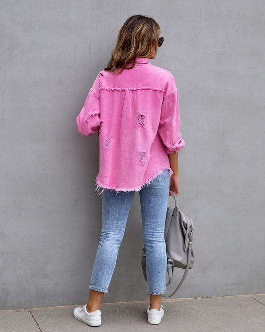 Woman wearing pink ripped shirt jacket with rolled sleeves, light blue jeans, and white sneakers, holding gray bag, standing against gray wall