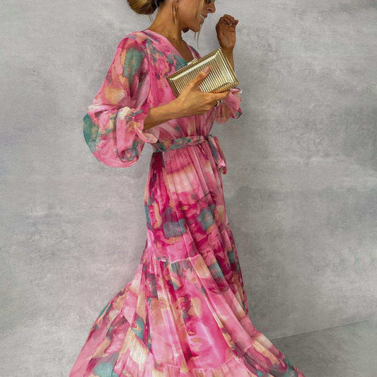 Woman wearing pink long-sleeved floral print chiffon maxi dress with V-neck and pleated high waist, holding gold clutch.