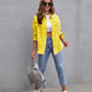 Woman wearing yellow ripped shirt jacket with ripped blue jeans and white sneakers standing against gray wall