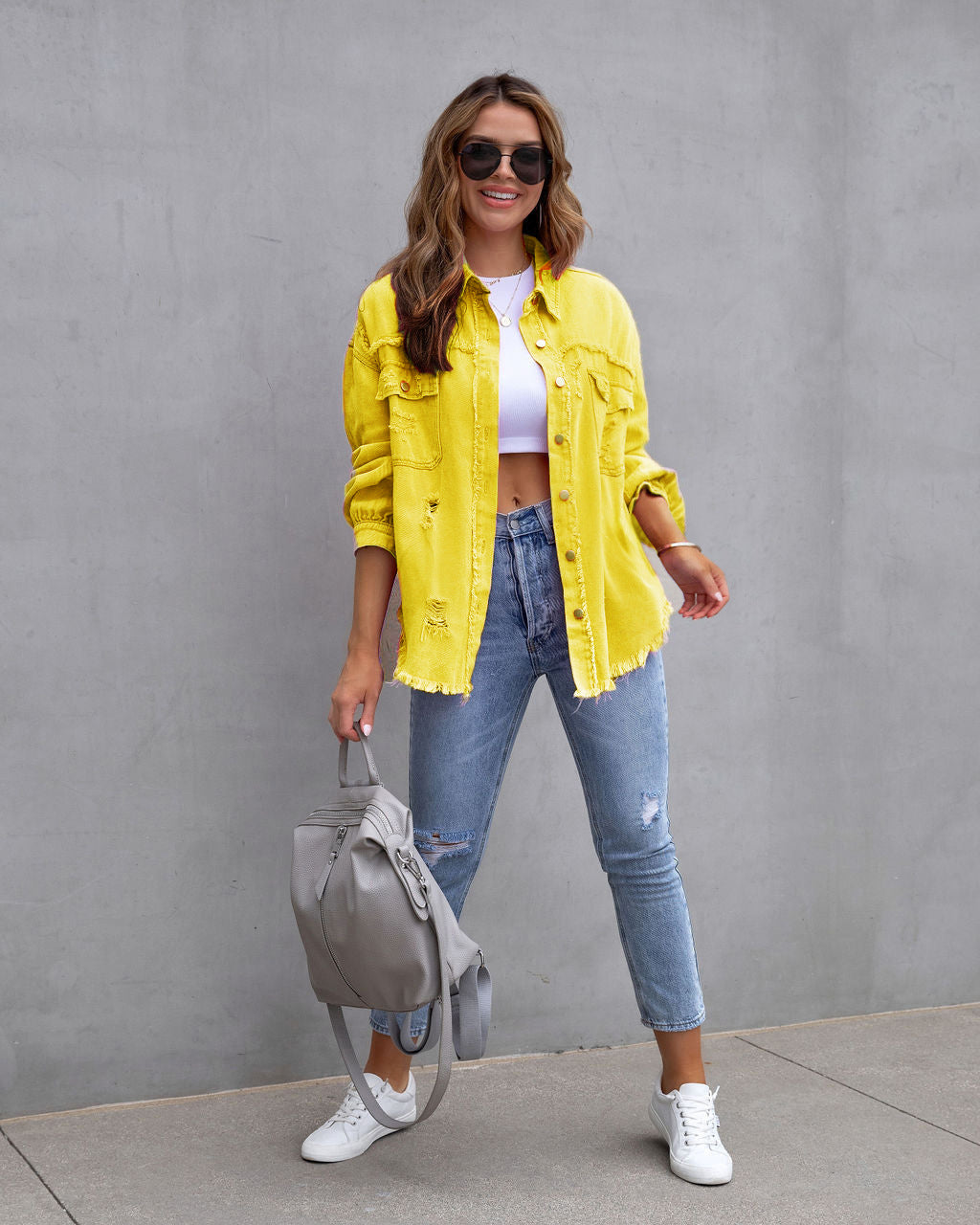 Woman wearing yellow ripped shirt jacket with ripped blue jeans and white sneakers standing against gray wall