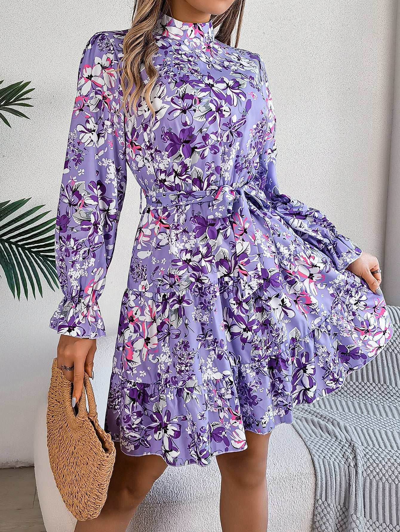 Woman wearing standing collar floral print long sleeve purple ruffled A-line dress with elastic waist and holding woven bag