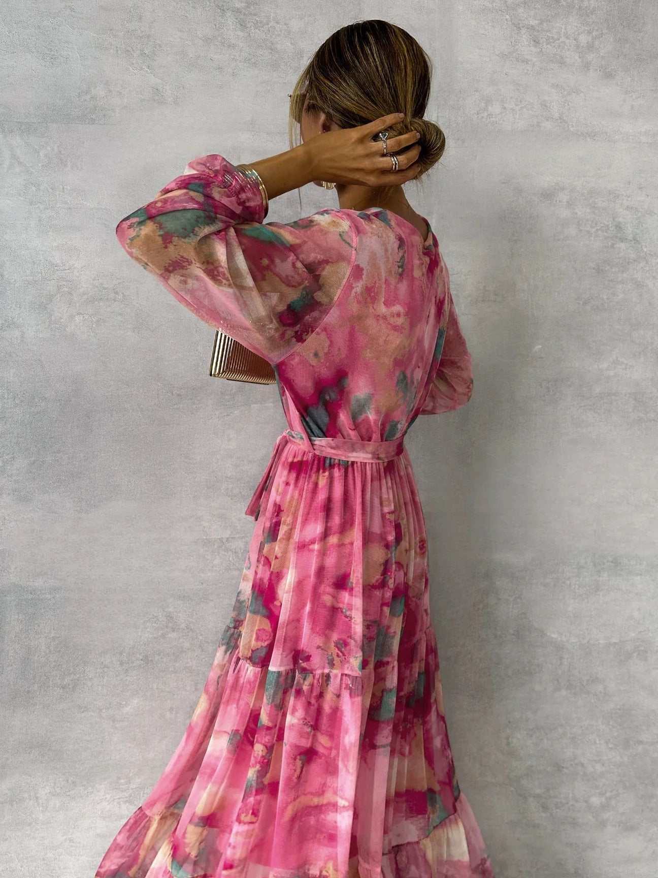 Woman wearing rose red chiffon long-sleeved floral print dress with ruffle sleeves and pleated high-waisted maxi skirt standing against gray wall