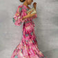 Woman wearing pink long-sleeved floral print chiffon maxi dress with V-neck and pleated high waist, holding gold clutch.