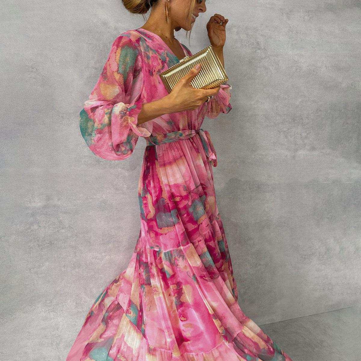 Woman wearing pink long-sleeved floral print chiffon maxi dress with V-neck and pleated high waist, holding gold clutch.